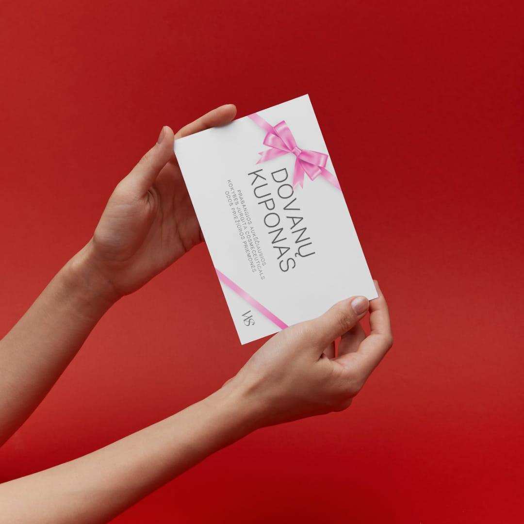 A pair of hands holding a Wellskino gift card against a bold red background. The white card features an elegant pink ribbon design and the text 'Dovanų Kuponas' (Gift Voucher) in Lithuanian.
