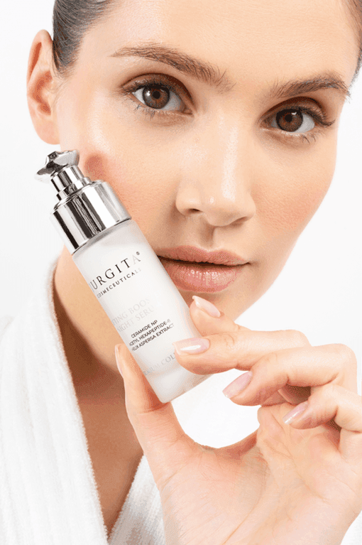 A close-up of a model with radiant skin gently holding the Jurgita Lifting Booster Night Serum bottle, showcasing its premium packaging.