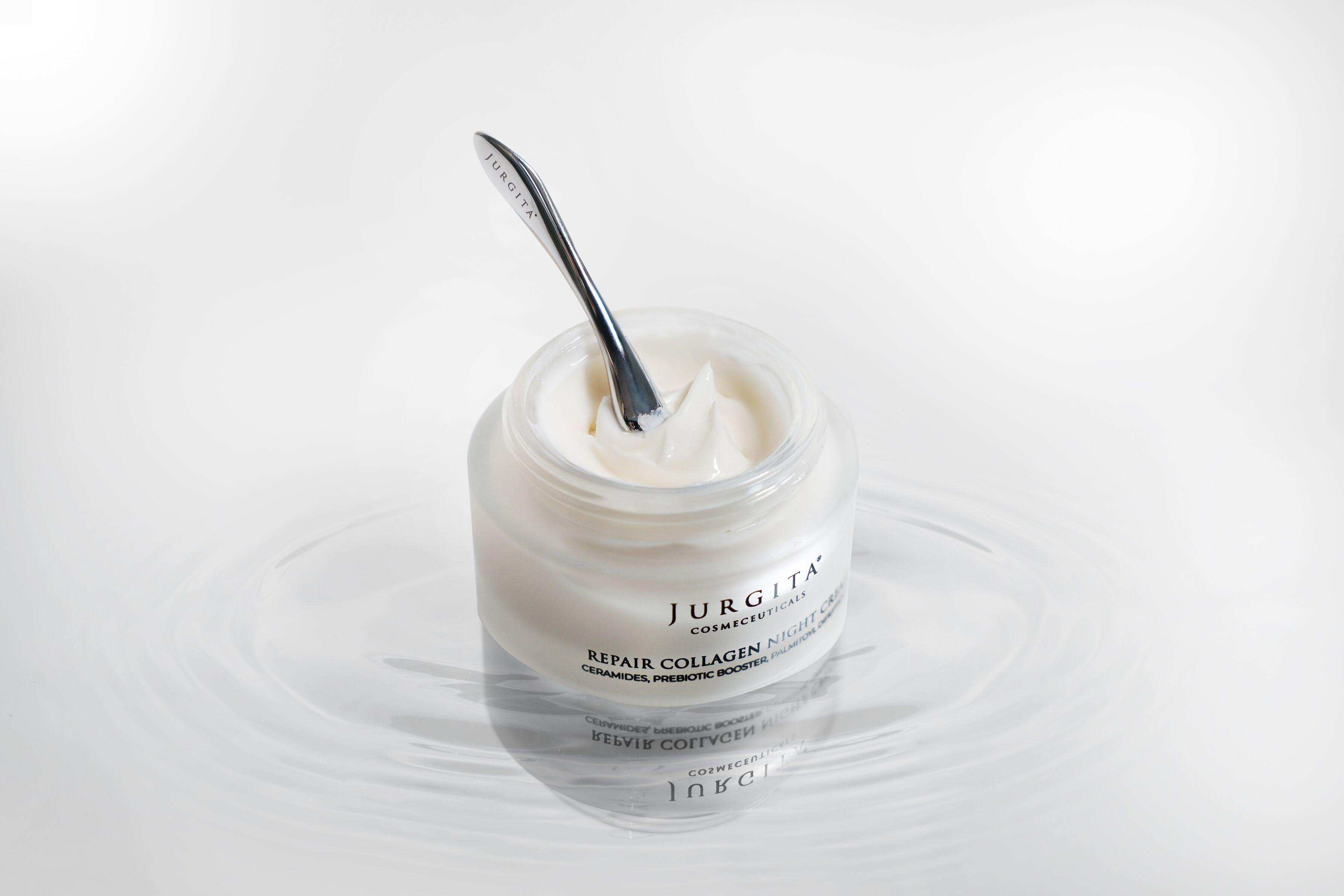 An open jar of Repair Collagen Night Cream with a sleek silver spatula, sitting on a reflective surface, emphasizing its rich and hydrating formula.