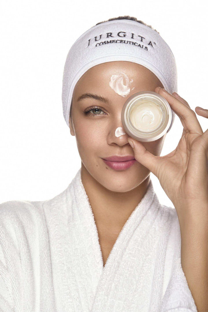 A woman in a white spa headband holding the Repair Collagen Night Cream jar up to her eye, with a soft application of cream on her face, showcasing its luxurious texture.